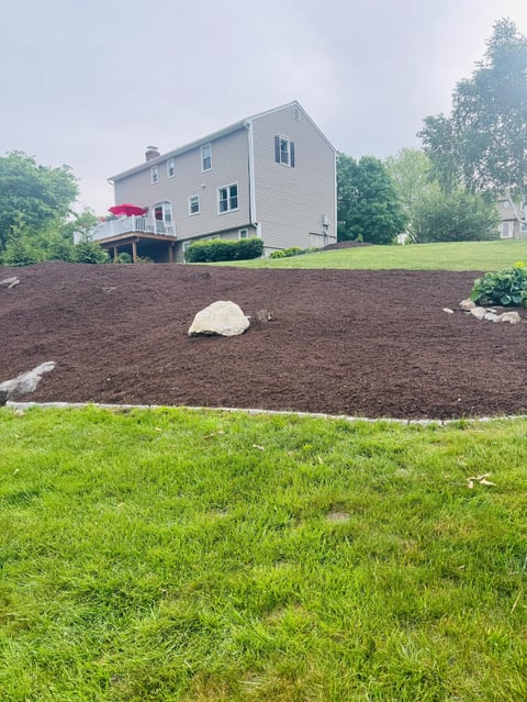 Two-story house with light gray siding on a grassy hillside with freshly mulched landscaping beds and decorative rocks