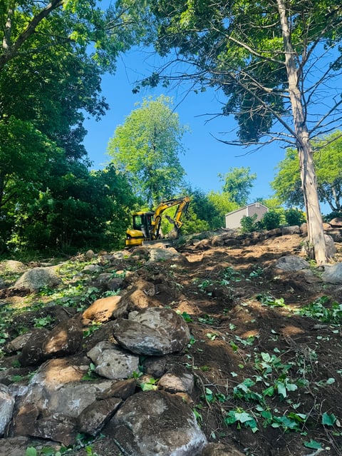 Construction excavator clearing rocky terrain surrounded by trees under blue sky